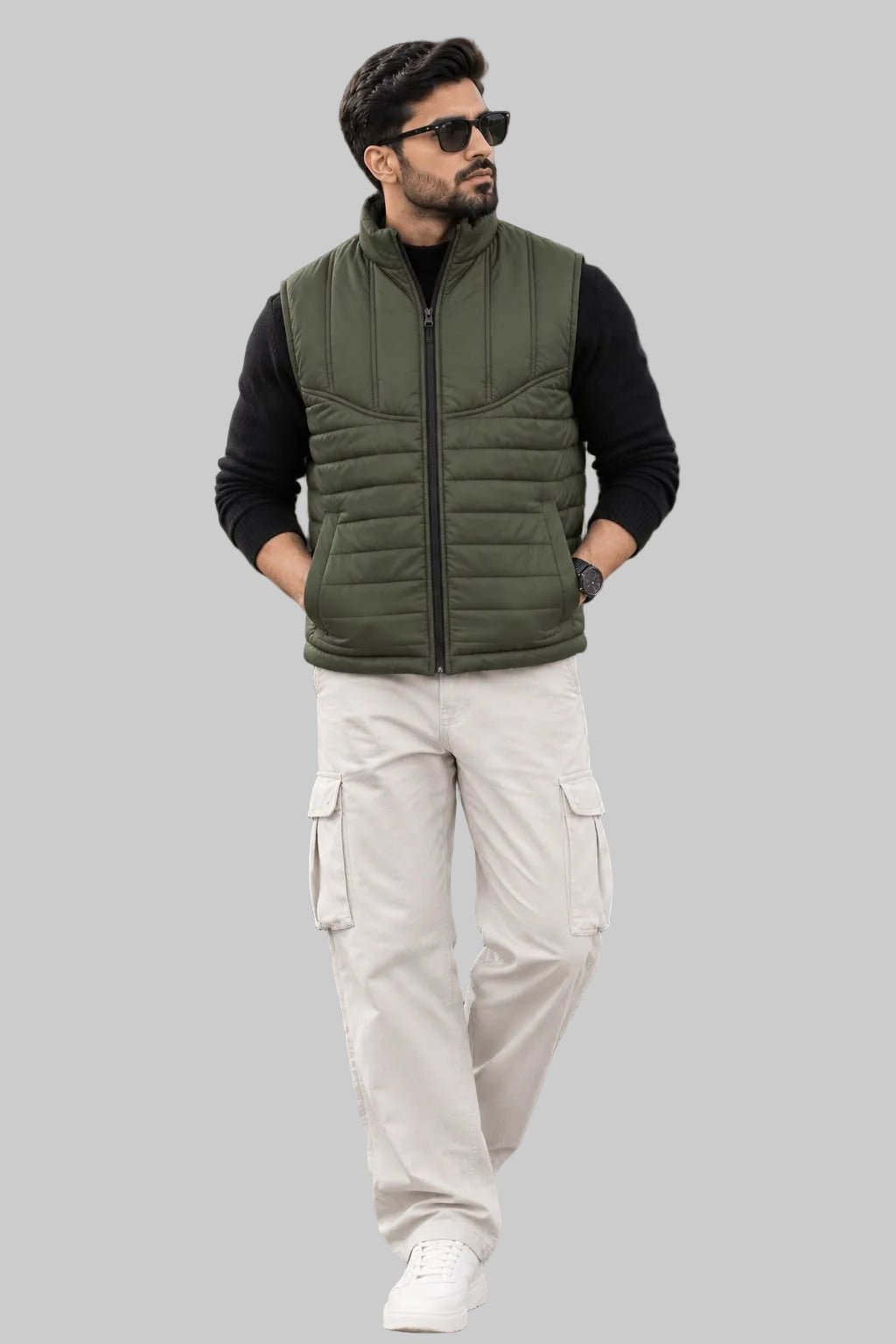A man wearing a sleeveless olive green puffer vest over a black long-sleeve shirt, cream cargo pants, and white sneakers, standing on a stone patio in front of a modern lodge with mountains in the background.
