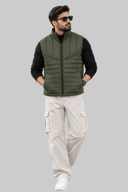 A man wearing a sleeveless olive green puffer vest over a black long-sleeve shirt, cream cargo pants, and white sneakers, standing on a stone patio in front of a modern lodge with mountains in the background.