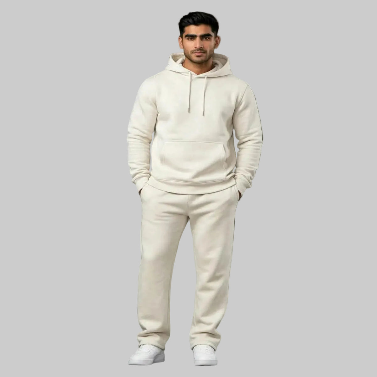 A man modeling a matching cream-colored hoodie and sweatpants lounge set against a dark gray textured studio backdrop.