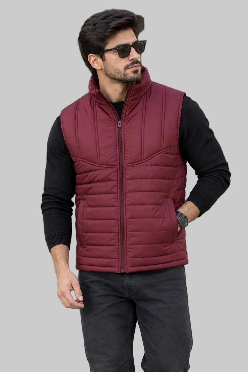 A man wearing a burgundy quilted zip-up puffer vest over a black long-sleeve shirt and dark gray jeans, standing outdoors in front of a modern building.