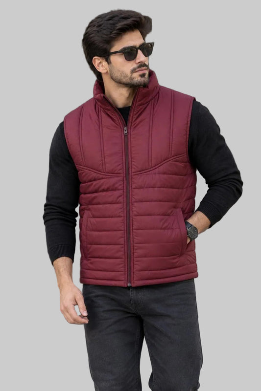 A man wearing a burgundy quilted zip-up puffer vest over a black long-sleeve shirt and dark gray jeans, standing outdoors in front of a modern building.